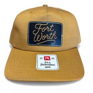 May include: A tan baseball cap with a flat brim. The cap features a black rectangular patch with "Fort Worth" embroidered in gold script. A white tag with the Richardson logo is on the brim.