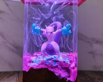 May include: A wooden-framed night light featuring a purple and white Mewtwo figure inside a clear resin block. The resin is illuminated with purple and blue lighting, creating a glowing effect. The base and top are made of wood.