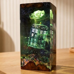 May include: A rectangular resin lamp featuring a dark, swirling green nebula effect with a miniature blue police box embedded inside. The base is made of natural wood, and the structure glows with an internal light, creating a deep, immersive scene.