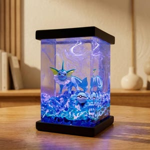 May include: A rectangular resin lamp featuring a Vaporeon figure submerged in a blue, swirling liquid environment. The base and top are black, and the interior is illuminated with blue light, showcasing the character and a small Poké Ball sphere.
