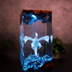 May include: A rectangular resin art piece featuring a white and blue mythical creature figurine. The figurine is set within a clear resin block with blue lighting effects and a dark wood top. The piece has a unique, artistic design.