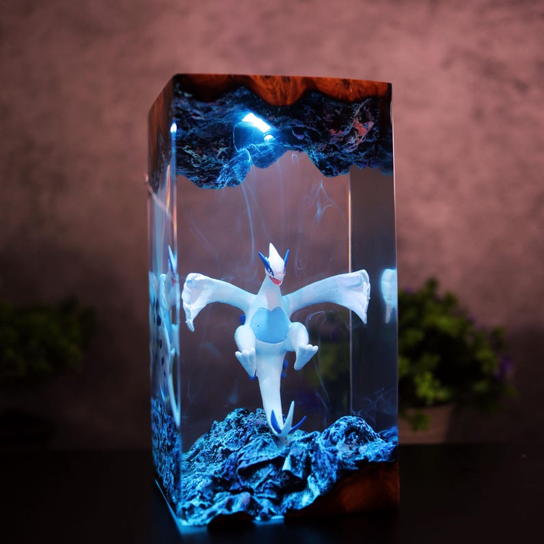 May include: A rectangular resin art piece featuring a white and blue mythical creature figurine. The figurine is set within a clear resin block with blue lighting effects and a dark wood top. The piece has a unique, artistic design.