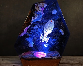 May include: A unique resin art piece featuring a glowing spaceship design within a blue and purple galaxy scene. The artwork is encased in a hexagonal shape with a wooden base, creating a striking decorative item.