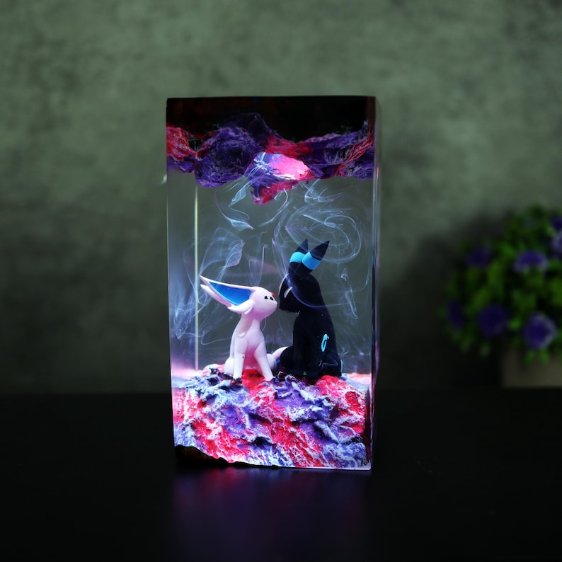 May include: A rectangular resin sculpture featuring two stylized figures, one white and one black, set against a backdrop of swirling white and purple hues. The base is a textured mix of red, purple, and blue.