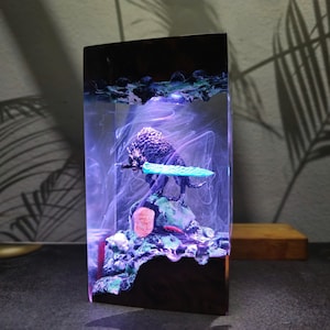 Può includere: Scultura rettangolare in resina raffigurante una creatura simile a un lupo che tiene in mano un oggetto turchese, posta su una base rocciosa. La scultura è racchiusa in resina trasparente con accenti bianchi e blu a spirale.