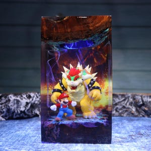 Pu&ograve; includere: Un blocco rettangolare in resina contenente le statuette di Bowser e Mario. La scena presenta un bordo superiore in legno naturale, uno sfondo luminoso nei toni del viola e dell'arancione che ricorda il fuoco e una base rocciosa colorata.