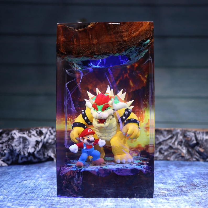 Pu&ograve; includere: Un blocco rettangolare in resina contenente le statuette di Bowser e Mario. La scena presenta un bordo superiore in legno naturale, uno sfondo luminoso nei toni del viola e dell'arancione che ricorda il fuoco e una base rocciosa colorata.