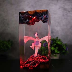 May include: A clear resin block contains a white and purple figurine, lit by red light. The top has a dark, textured design. The figurine rests on a red, rocky base, forming a unique decorative item. The piece is approximately 15 cm tall.