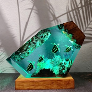 May include: A decorative resin sculpture featuring a vibrant underwater scene with sea turtles. The clear resin is illuminated with a turquoise glow, showcasing the turtles and coral. The sculpture is set on a wooden base, with a dark wood and bark element.