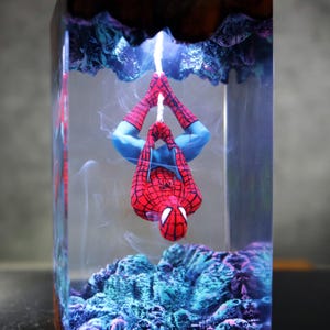 Puede incluir: Una figura de Spider-Man roja y azul colgando boca abajo dentro de un bloque de resina transparente. El bloque presenta formaciones rocosas azules y moradas en la parte superior e inferior. La figura cuelga de una cuerda blanca.