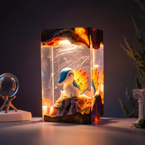 Peut inclure: Lampe rectangulaire avec un personnage Pokémon dans de la résine transparente. La lampe dégage une lumière chaude, mettant en valeur les couleurs bleu, blanc et orange du personnage. La résine est encadrée par un bord effet bois.