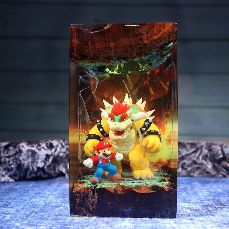 Pu&ograve; includere: Un blocco rettangolare in resina contenente un set di statuette di Bowser e Mario. La scena presenta una sezione superiore in legno scuro, un'area centrale con uno sfondo arancione e giallo infuocato e una base testurizzata al centro.