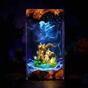May include: A rectangular resin and wood block lamp featuring a Mimikyu figure nestled among sculpted trees and green foliage. The interior is illuminated with a blue light, creating a glowing effect that highlights the character and the surrounding landscape.