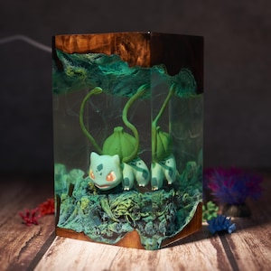 May include: A rectangular wooden block with a Bulbasaur figure encased in clear resin. The resin has a green and blue hue, with a wood grain top. The Bulbasaur is light blue with red eyes and green bulb. Decorative item.