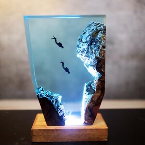 Könnte beinhalten: Harzskulptur mit zwei Tauchern in einer blauen Unterwasserlandschaft. Das Kunstwerk umfasst Felsformationen und ist auf einem Holzsockel montiert. Das Stück wird von einem sanften, blauen Licht beleuchtet.