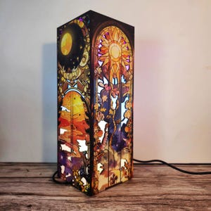 May include: A rectangular lamp with a stained-glass design featuring celestial and floral motifs. The artwork depicts a sun, moon, and stars in vibrant colors. The lamp is illuminated, showcasing the intricate details of the design. A black power cord extends from the base.