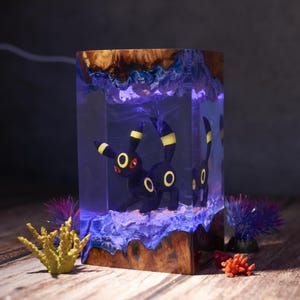 Może przedstawiać: Prostokątna drewniana lampa z żywicznym wzorem. Lampa przedstawia czarną figurkę Umbreona z żółtymi akcentami wewnątrz przezroczystego bloku z żywicy. Żywica ma niebiesko-fioletową poświatę, z drewnianą podstawą i górą. Ozdobne koralowce i rośliny na pierwszym planie.