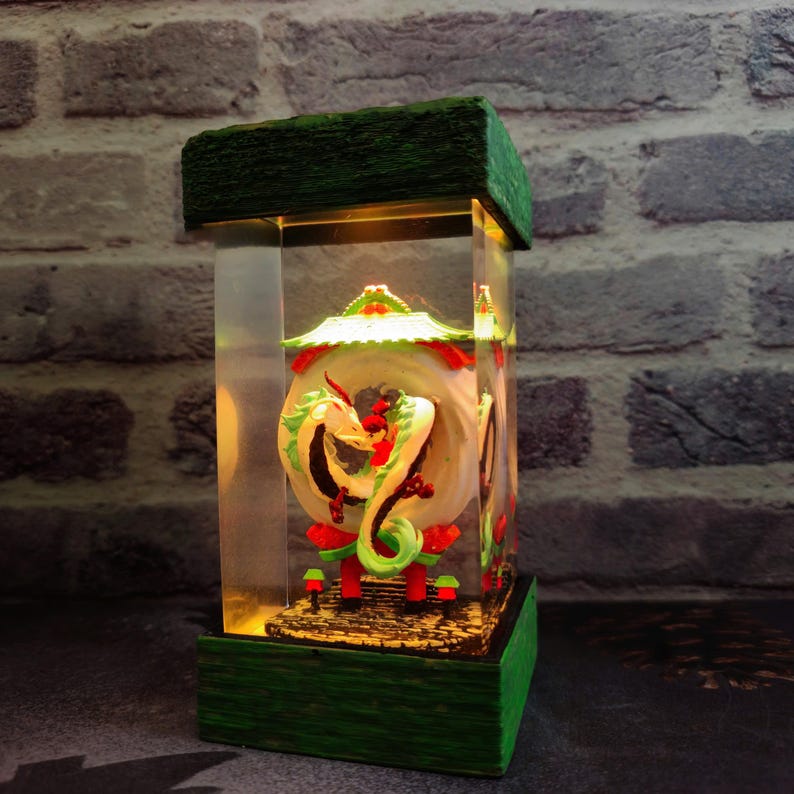 Op de afbeelding: Een decoratieve, rechthoekige lamp met een groene basis en bovenkant. Binnenin is een gedetailleerde sculptuur van een groene en witte draak te zien, tegen een achtergrond van een cirkelvormig ontwerp. De lamp heeft een warme gloed.