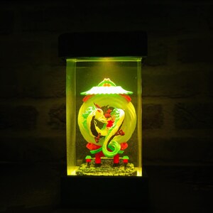 Op de afbeelding: Een rechthoekige, verlichte display case met een groene en gele drakenbeeldhouwwerk. De draak is opgerold rond een gele bol, met rode en groene accenten. De case heeft een zwarte basis en bovenkant, en de binnenkant wordt verlicht met een warm geel licht.