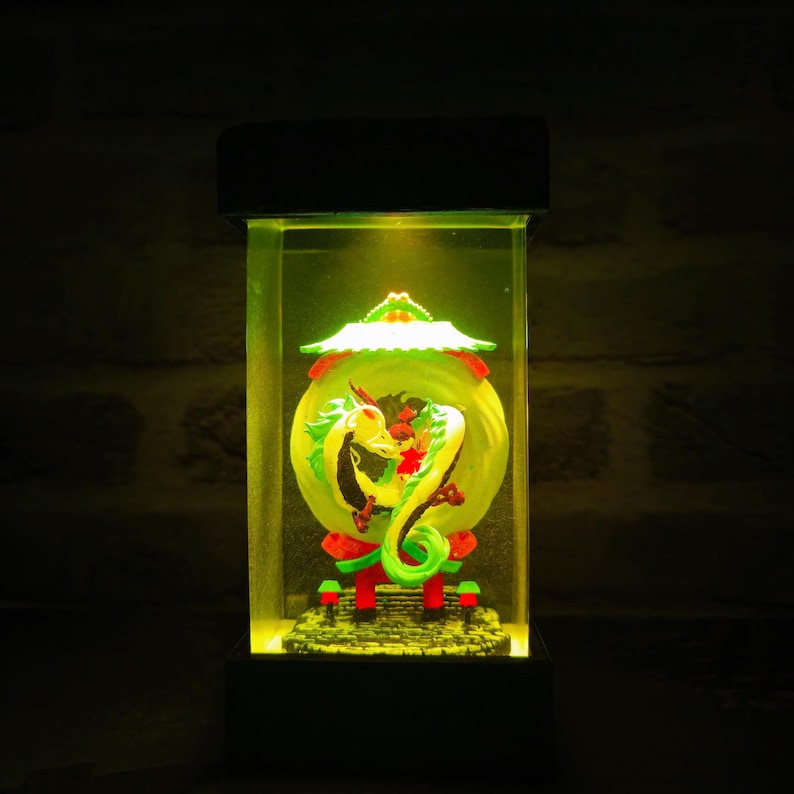 Op de afbeelding: Een rechthoekige, verlichte display case met een groene en gele drakenbeeldhouwwerk. De draak is opgerold rond een gele bol, met rode en groene accenten. De case heeft een zwarte basis en bovenkant, en de binnenkant wordt verlicht met een warm geel licht.