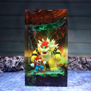 Pu&ograve; includere: Un blocco rettangolare in resina contenente le statuette di Bowser e Mario. La parte superiore &egrave; in legno naturale scuro, mentre l'interno presenta uno sfondo infuocato arancione e giallo con striature verdi che circondano i due personaggi.