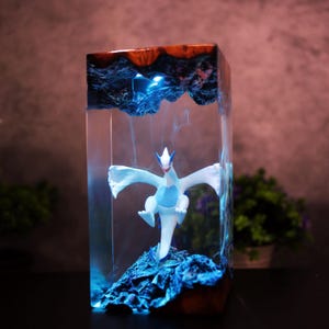 May include: A rectangular resin sculpture featuring a white and blue mythical creature figure. The sculpture has a blue and brown top, with blue lighting effects. The figure is standing on a blue rock formation inside the clear resin.