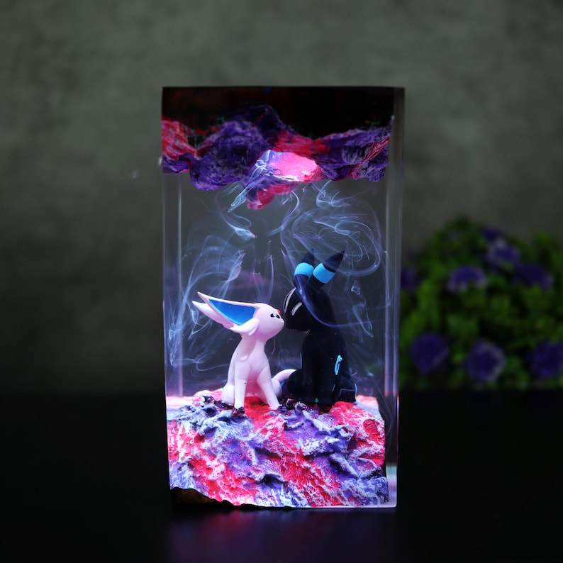 May include: A resin sculpture featuring two stylized figures, one pink and one black, set on a colorful base. The figures are enclosed in a clear rectangular block with swirling, smoky effects and a vibrant, layered top.
