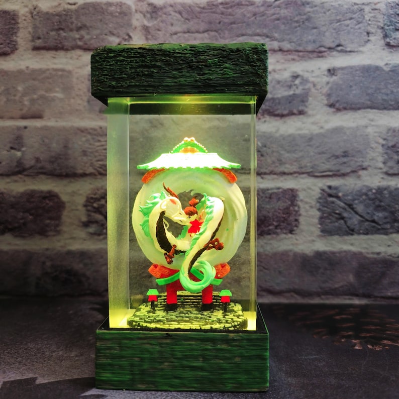 Op de afbeelding: Een decoratieve, verlichte display met een drakenbeeld. De draak is wit en groen, met rode accenten, tegen een cirkelvormige achtergrond. De basis en de bovenkant zijn groen en het licht schijnt van binnenuit.