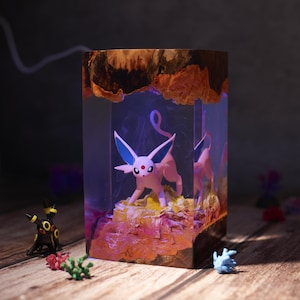 Op de afbeelding: Decoratief harsblok met een roze stripfiguur met blauwe oren, tegen een kleurrijke, verlichte achtergrond. Het blok heeft een houten boven- en onderkant en het figuur is omgeven door een levendig, gestructureerd landschap.