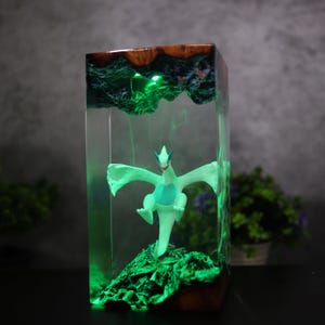 May include: A rectangular resin art piece featuring a white and teal mythical creature figurine. The figurine is set on a green, textured base within a clear resin block. The top is a mix of wood and green resin, with a green light illuminating the piece.