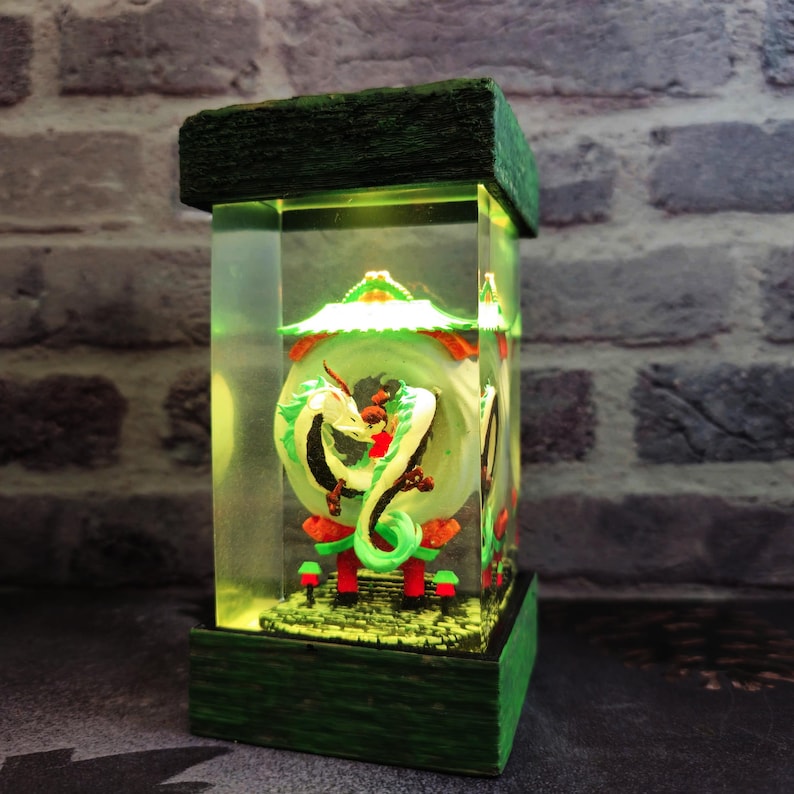 Op de afbeelding: Een rechthoekige, verlichte decoratieve lamp met een groen en wit drakenontwerp. De lamp heeft een groene basis en bovenkant, met een heldere, rechthoekige behuizing. De draak staat tegen een groene achtergrond met rode en oranje accenten.