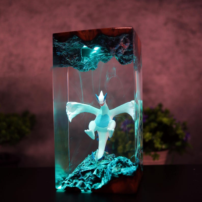 May include: A rectangular resin art piece featuring a white and blue mythical creature figurine. The figurine is set within a clear resin block with a teal-coloured, textured base and top. The wooden top adds a natural contrast to the piece.