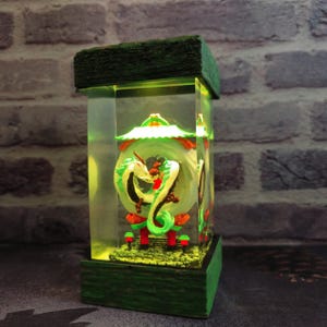 Op de afbeelding: Een rechthoekige, verlichte decoratieve lamp met een groen en wit drakenontwerp. De lamp heeft een groene houten basis en bovenkant, met een heldere, rechthoekige behuizing. De draak staat tegen een groene achtergrond met rode en oranje accenten.