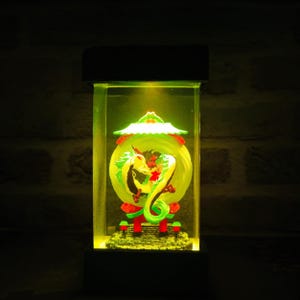 Op de afbeelding: Een decoratieve verlichte doos met een drakenbeeld. De draak is geel, groen en rood, tegen een gele achtergrond. De doos heeft een zwarte basis en bovenkant, met helder licht van bovenaf.