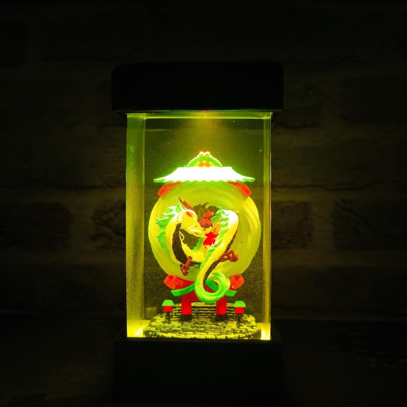Op de afbeelding: Een decoratieve verlichte doos met een drakenbeeld. De draak is geel, groen en rood, tegen een gele achtergrond. De doos heeft een zwarte basis en bovenkant, met helder licht van bovenaf.