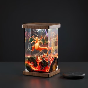 Puede incluir: Lámpara rectangular con una figura de Charizard dentro de un bloque de resina transparente. La lámpara tiene una base y una parte superior de madera. La escena incluye efectos de lava y humo, con un fondo negro y un pequeño objeto negro redondo.