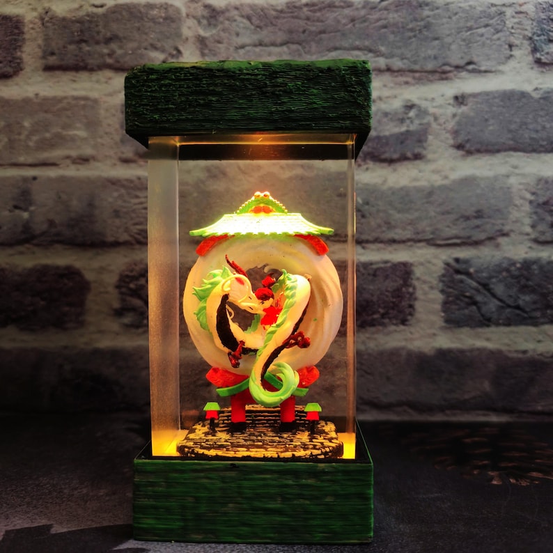 Op de afbeelding: Een decoratieve, verlichte lantaarn met een gedetailleerde drakenbeeldhouwkunst. De draak is groen, rood en wit, tegen een cirkelvormige achtergrond. De lantaarn heeft een rechthoekige vorm met een groene basis en bovenkant en een helder middenstuk.