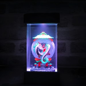 Op de afbeelding: Een rechthoekige, verlichte displaycase met een gedetailleerde sculptuur van een witte en turquoise draak die een rode figuur omarmt in een bol. De sculptuur staat tegen een blauwe achtergrond, met een rood en groen dak in pagodestijl.