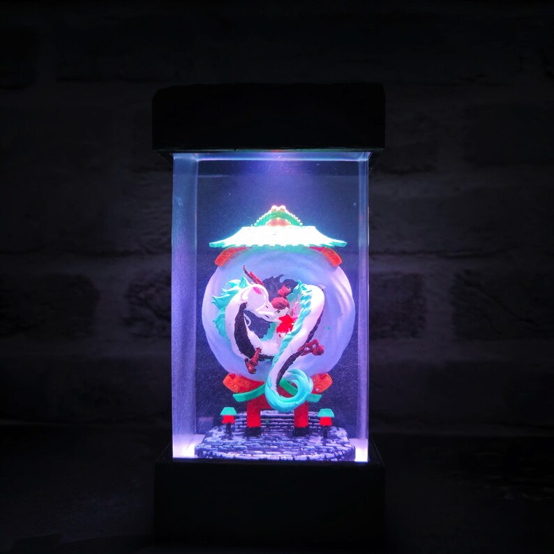 Op de afbeelding: Een rechthoekige, verlichte displaycase met een gedetailleerde sculptuur van een witte en turquoise draak die een rode figuur omarmt in een bol. De sculptuur staat tegen een blauwe achtergrond, met een rood en groen dak in pagodestijl.