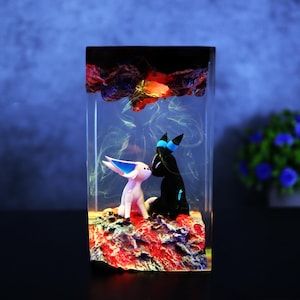 May include: A rectangular resin art piece featuring two cartoon figures, one white with blue accents and the other black with blue accents, set on a red and brown base. The top has a dark, textured design with orange and yellow highlights.