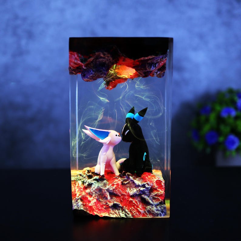 May include: A rectangular resin art piece featuring two cartoon figures, one white with blue accents and the other black with blue accents, set on a red and brown base. The top has a dark, textured design with orange and yellow highlights.