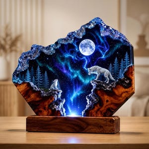 May include: A decorative lamp featuring a resin and wood sculpture of two wolves in a forest under a full moon and aurora borealis. The scene is set within a jagged, crystal-like frame, mounted on a rectangular wooden base with integrated lighting.