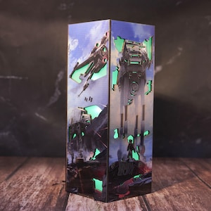 May include: A rectangular wooden lamp featuring a Star Wars theme. The lamp has cut-out designs that allow green light to shine through, depicting scenes with spaceships and AT-AT walkers against a blue sky and rocky terrain background. The lamp is on a wooden surface.