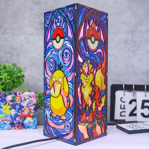 May include: A rectangular lamp featuring stained glass-style panels with illustrations of Psyduck and Arcanine. The design includes blue swirling patterns, red and white Poke Ball motifs, and dark wooden edges. The lamp is illuminated from within.