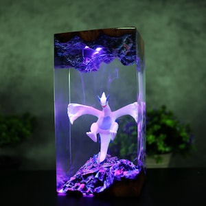 May include: A rectangular resin art piece featuring a white and blue mythical creature figurine. The figurine is set against a backdrop of purple-lit rock formations and encased in clear resin with a wooden top.