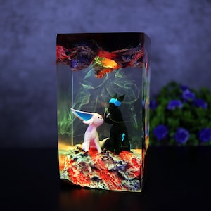 May include: A resin art piece featuring two cartoon figures inside a clear rectangular block. One figure is white with blue accents, the other is black with blue accents. The base is red and the top has colorful swirls.