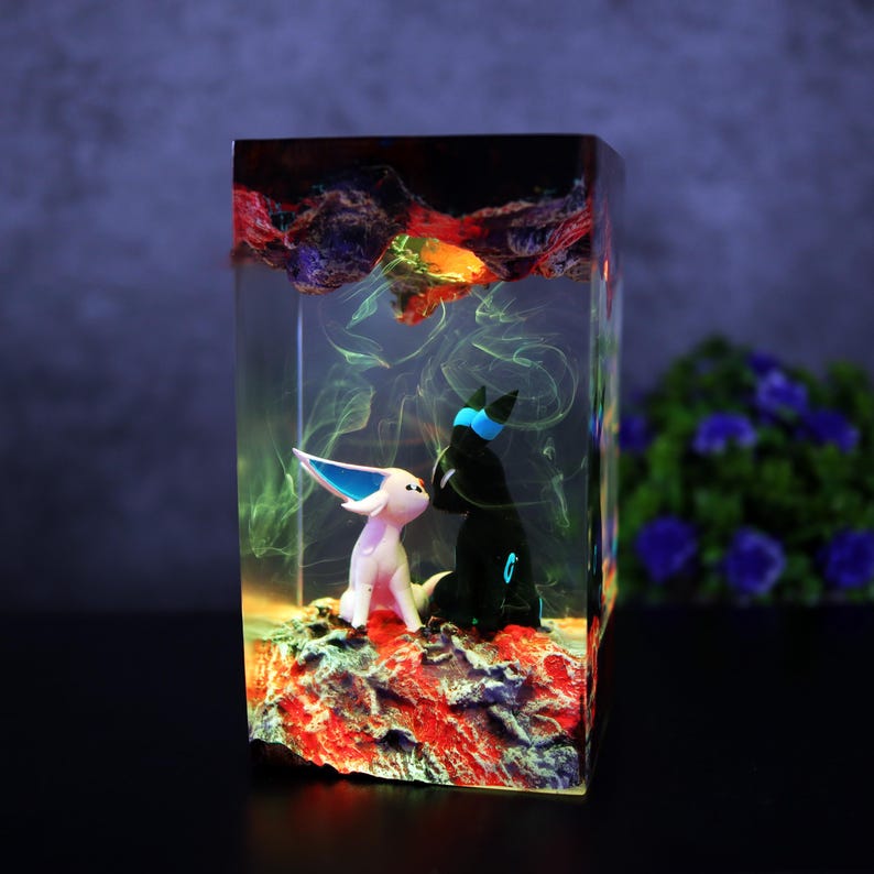 May include: A resin art piece featuring two cartoon figures inside a clear rectangular block. One figure is white with blue accents, the other is black with blue accents. The base is red and the top has colorful swirls.