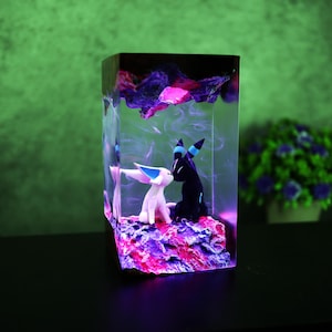 May include: A rectangular resin sculpture featuring two stylized animal figures in a loving embrace. The sculpture has a vibrant color palette of pink, purple, and blue, with a glowing effect. The background is a gradient of green.