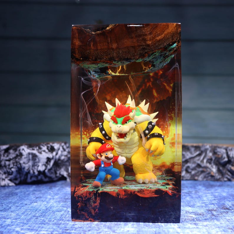 Pu&ograve; includere: Un blocco rettangolare in resina contenente una statuetta di Bowser e una di Mario. La scena presenta uno sfondo arancione e giallo infuocato con una sezione superiore in legno scuro. Le figure sono posizionate su una base testurizzata.