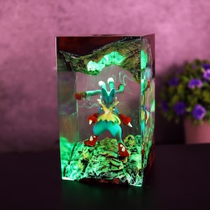 Puede incluir: Una pieza de arte de resina rectangular con una figura colorida de Lucario de pie sobre una base verde texturizada. La resina es transparente, con un efecto de brillo verde. La parte superior tiene un diseño orgánico marrón oscuro.